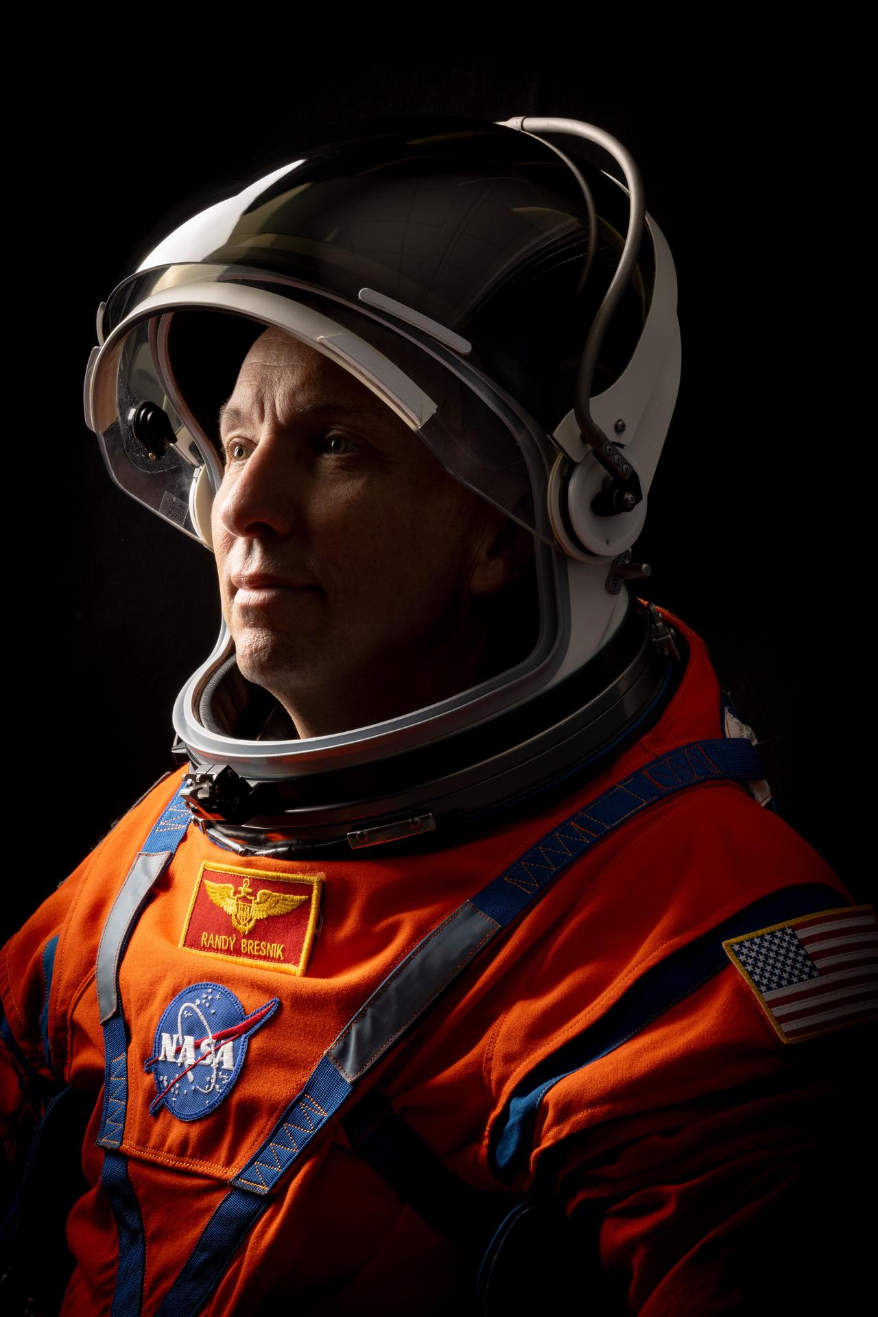 NASA astronaut Randy Bresnik is pictured in a close-up, side profile shot, wearing an orange Orion Crew Survival System suit with the helmet visor up and his face exposed. 