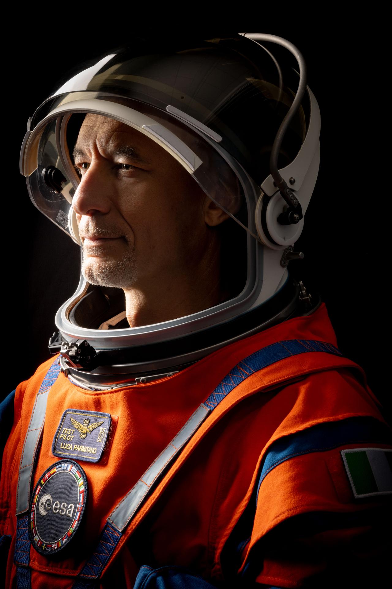 ESA (European Space Agency) astronaut Luca Parmitano is pictured in a close-up, side profile shot, wearing an orange Orion Crew Survival System suit with the helmet visor up and his face exposed. 
