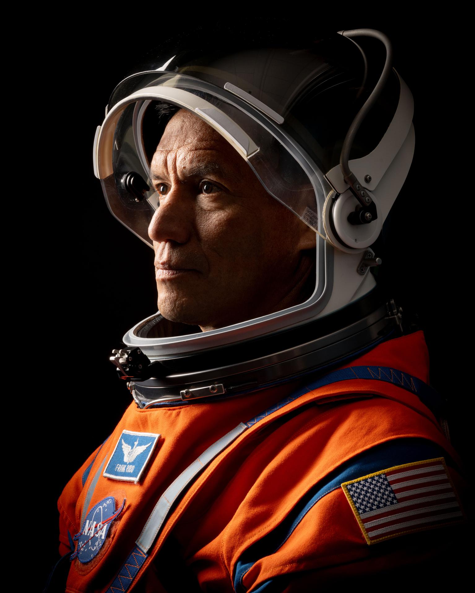 NASA astronaut Frank Rubio is pictured in a close-up, side profile shot, wearing an orange Orion Crew Survival System suit with the helmet visor up and his face exposed.