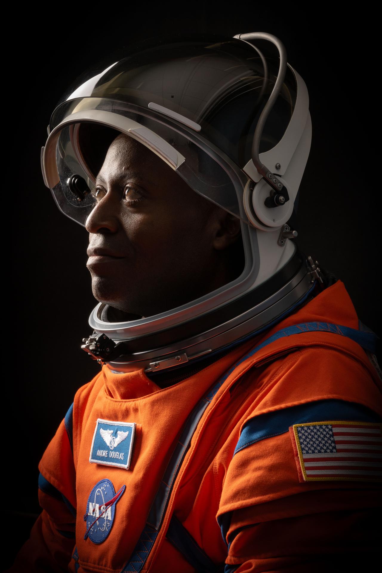 NASA astronaut Andre Douglas is pictured in a close-up, side profile shot, wearing an orange Orion Crew Survival System suit with the helmet visor up and his face exposed.