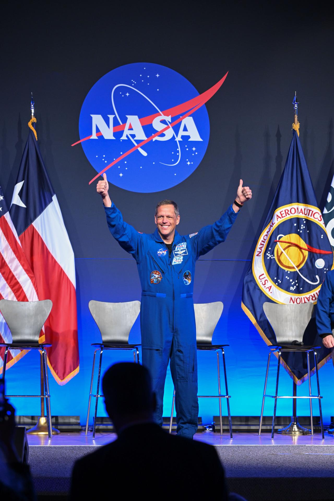 NASA astronaut Bob Hines is pictured wearing his blue flight suit with his arms raised, smiling. He is on-stage in front of four chairs. Behind the chairs is the NASA meatball, with American and Texas flags to the left and NASA and ESA (European Space Agency) flags to the right.