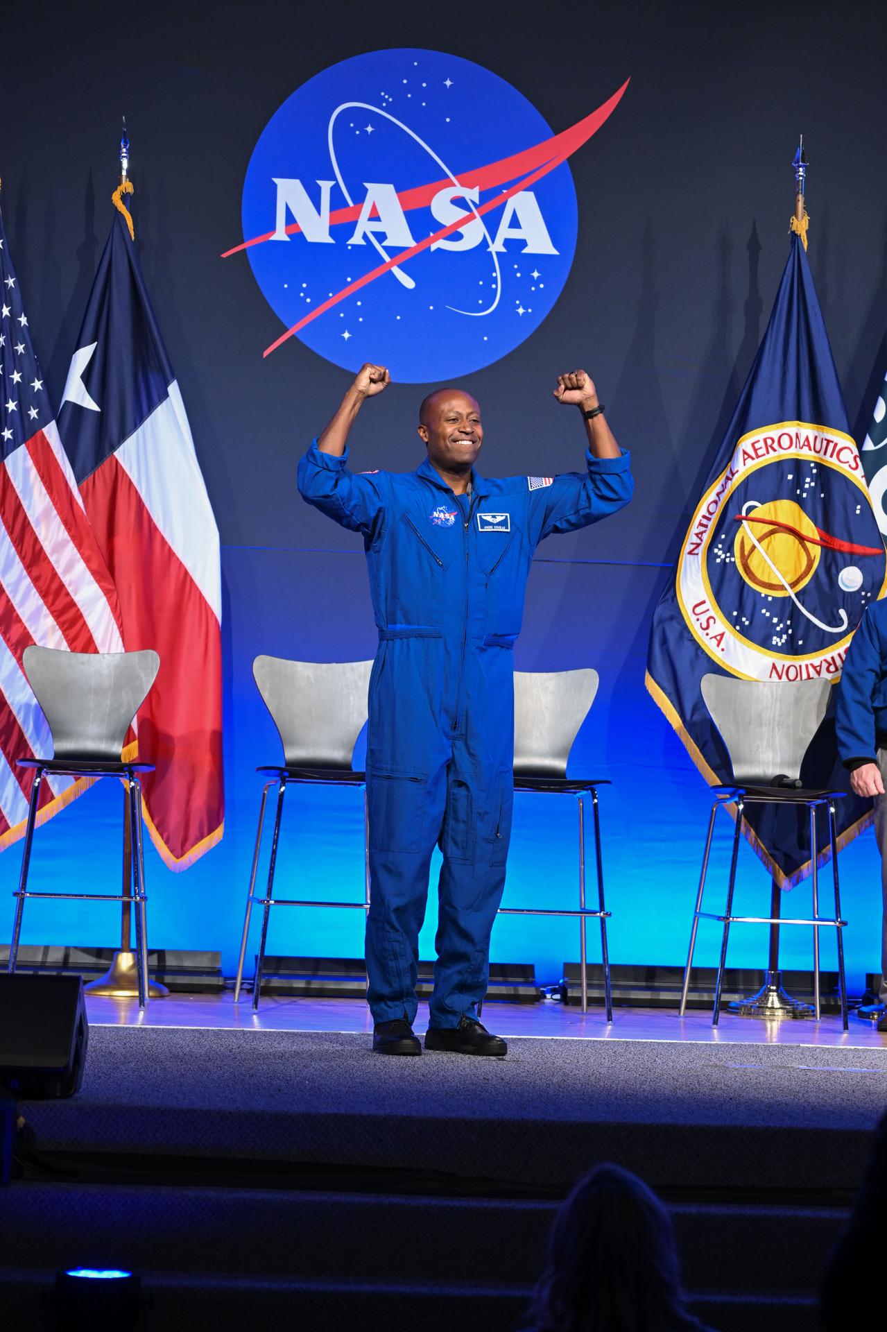 NASA astronaut Andre Douglas is pictured wearing his blue flight suit with his arms raised, smiling. He is on-stage in front of four chairs. Behind the chairs is the NASA meatball, with American and Texas flags to the left and NASA and ESA (European Space Agency) flags to the right.