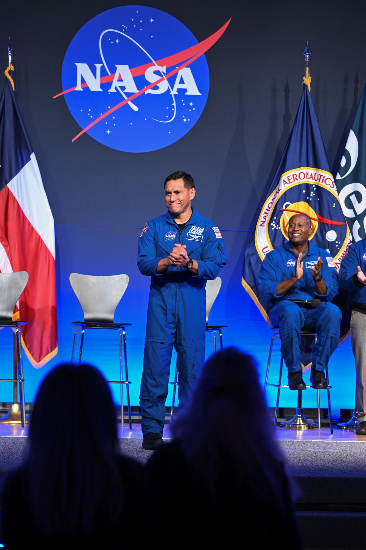 NASA astronaut Frank Rubio is pictured wearing his blue flight suit with his hands clasped in front of his chest, smiling. He is on-stage in front of four chairs. NASA astronaut Andre Douglas can be seen seated behind him, clapping and smiling. Behind the chairs is the NASA meatball, with the Texas flag to the left and NASA and ESA (European Space Agency) flags to the right.