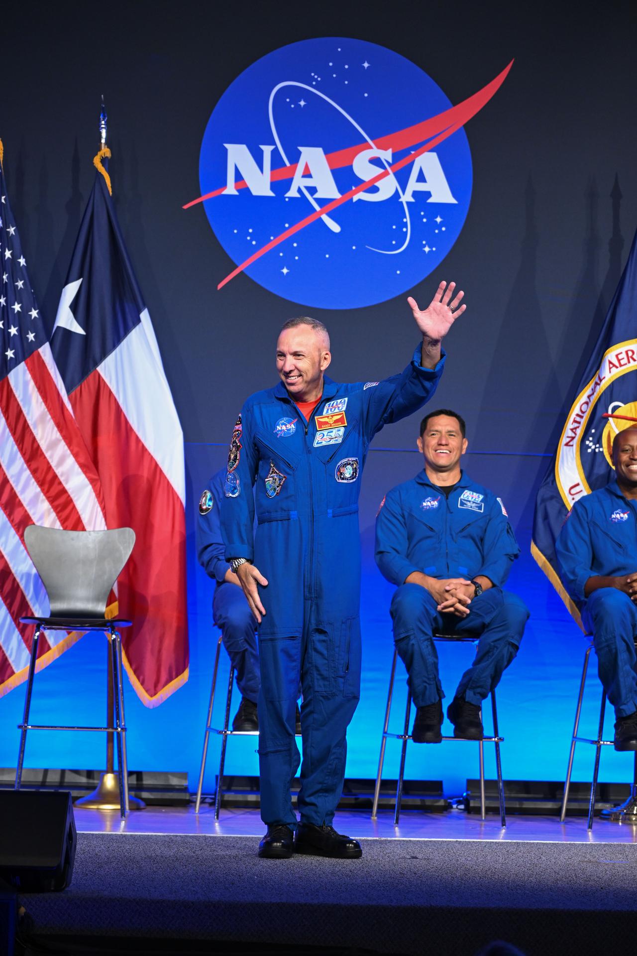 NASA astronaut Randy Bresnik is pictured wearing his blue flight suit with left arm raised, waving and smiling. He is on-stage in front of four chairs. NASA astronauts Frank Rubio and Andre Douglas can be seen seated behind him, smiling. Behind the chairs is the NASA meatball, with American and Texas flags to the left and a NASA flag to the right.