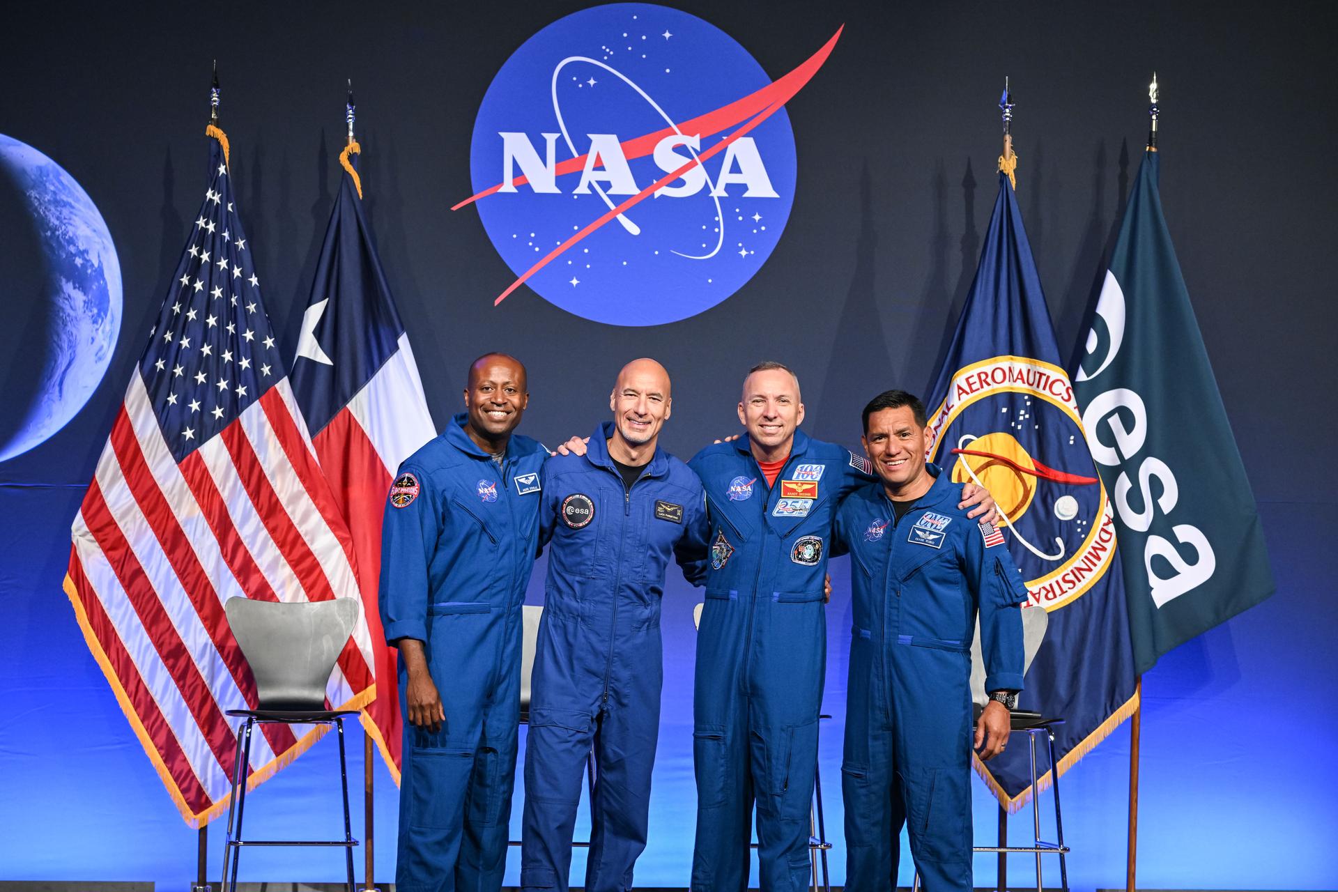 Four astronauts are pictured wearing their blue flight suits, standing with their arms around each other and smiling. They are on a stage with the NASA meatball behind them and American and Texas flags to the left and NASA and ESA (European Space Agency) flags to the right.