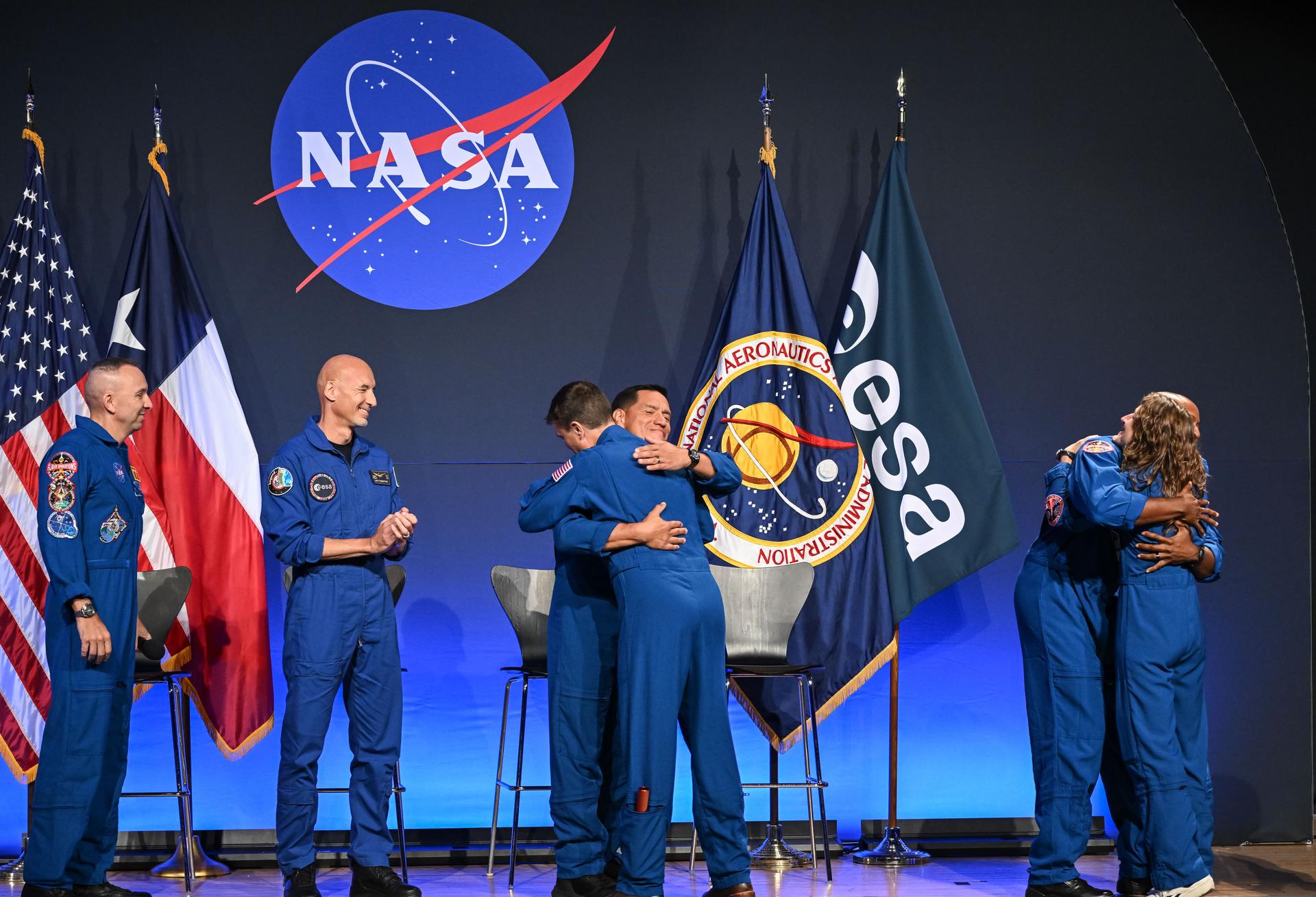 Six astronauts are pictured wearing blue flight suits. Two sets of two astronauts on the far right and center of the image are seen hugging, while the other two astronauts in the far left of the image look on, smiling. Behind all of the astronauts, the NASA meatball can be seen, with American and Texas flags pictured to the left and NASA and ESA (European Space Agency) flags to the right.