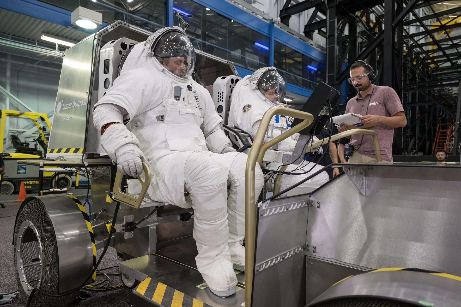 Artemis EVA training lead Scott Wray (left) and NASA astronaut Anne McClain (right) sit inside Lunar Outpost’s Pegasus lunar terrain vehicle prototype during human-in-the-loop testing at NASA’s Johnson Space Center where they evaluated the design, usability, and crew comfort of Lunar Outpost’s lunar rover. NASA selected Lunar Outpost and Astrolab in May 2026 to build and deliver the first phase of lunar terrain vehicles for Moon Base development. Image Credit: NASA/Robert Markowitz