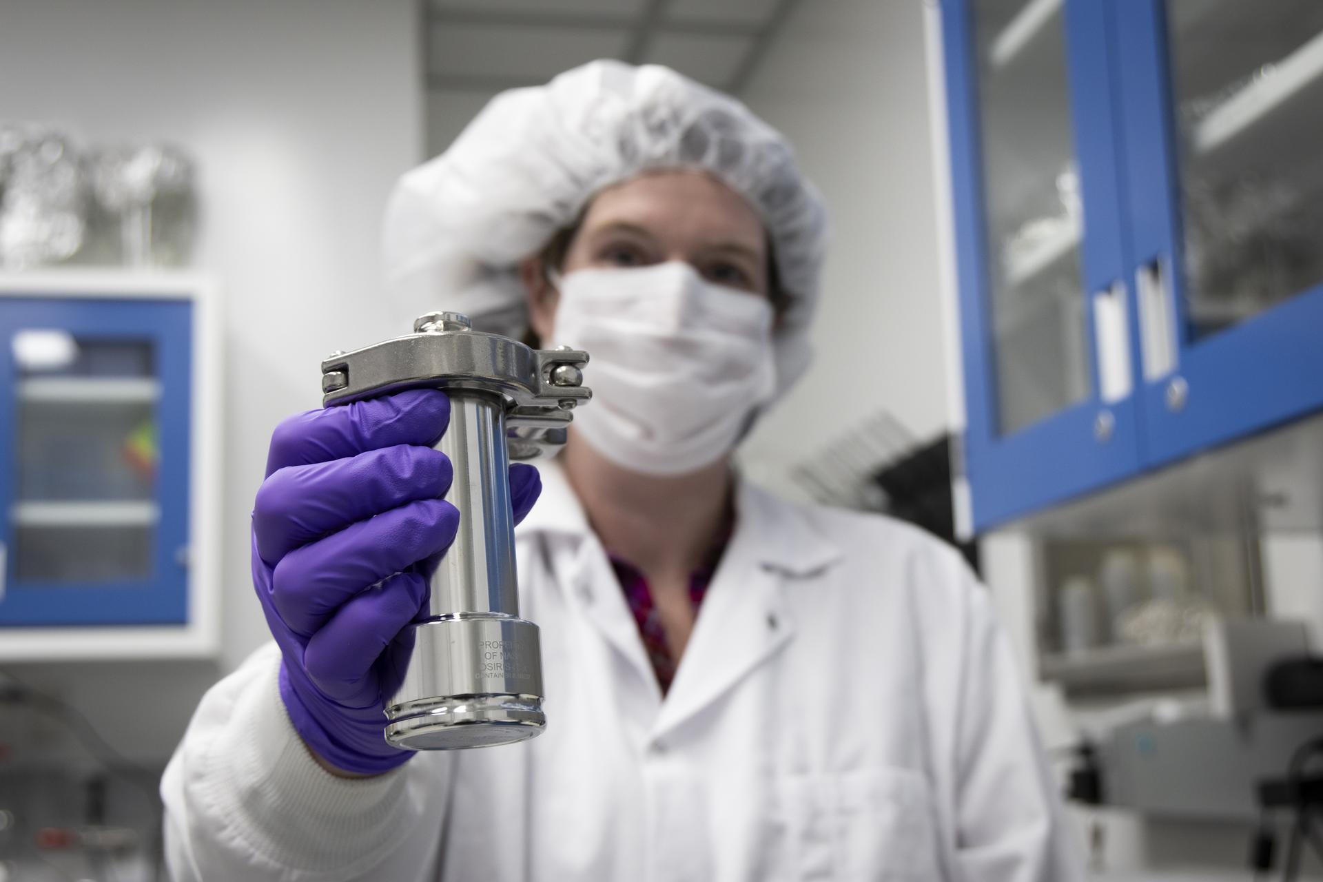 Hannah McLain stands inside an astrobiology lab, looking directly at the camera. She is dressed in a white lab coat, white surgical face mask, white bouffant cap, and purple nitrile gloves. Her arm extends towards the camera, presenting a cylindrical metal container that is slightly larger than her hand. The phrases “PROPERTY OF NASA,” “OSIRIS-REx,” and “CONTAINER #:90237” are engraved in small capital letters on the base.
