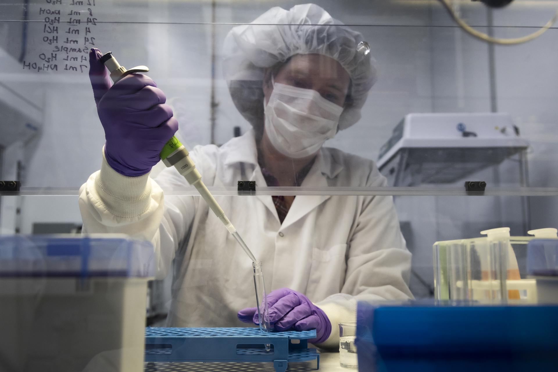 Hannah McLain sits inside an astrobiology lab, reaching under a fume hood. She is dressed in a white lab coat, white surgical face mask, white bouffant cap, and purple nitrile gloves. In her left hand, she holds a foot-long pipette containing a clear liquid. She transfers the pipette’s contents into a small test tube, which is seated in a blue test tube rack.