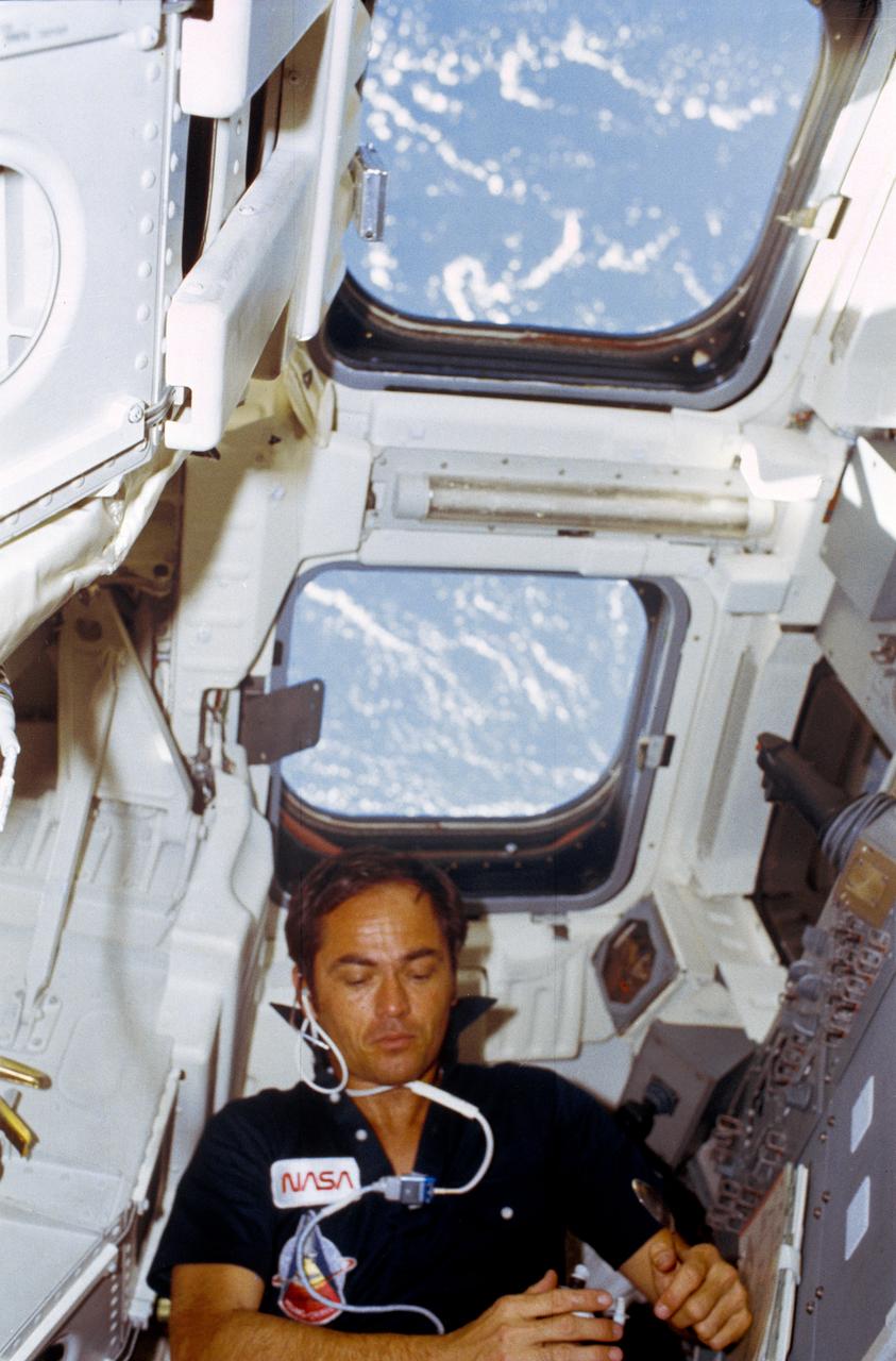 STS001-06-481 (April 1981) --- Astronaut Robert L. Crippen, STS-1 pilot, is behind his station in the flight deck of Columbia. He has a spoon in his hand. The windows prominent in the scene are the ones through which a number of Earth scenes were photographed with a 70mm camera by the two astronauts. Clouds over water can be seen through them. Just inches away from the top windows, not quite so obvious at top of the frame (if held horizontally) are the two aft cabin windows through which the crew viewed the cargo bay and the aft end of the craft, including the orbital maneuvering system (OMS) pods. Astronaut John W. Young, commander, took this photo with a 35mm camera. Photo credit: NASA