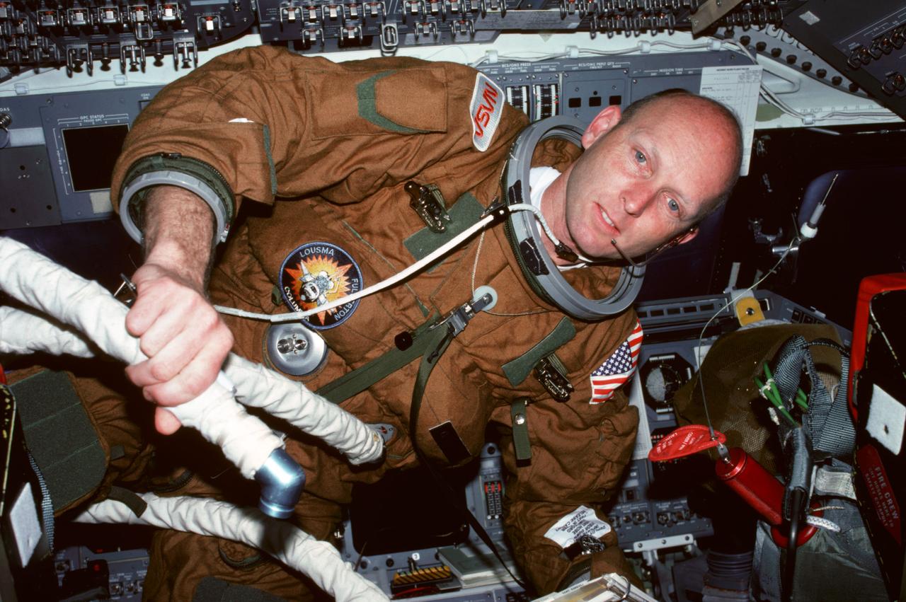 STS003-31-290 (30 March 1982) --- Astronaut Gordon Fullerton, STS-3 pilot, wearing communications kit assembly (ASSY) mini-headset (HDST) and ejection escape suit (EES), holds flexible hose attached to his EES vent hose fitting and second hose for commander's EES while behind pilots ejection seat (S2) seat back on the aft flight deck. Forward flight deck control panels are visible in the background. Photo credit: NASA