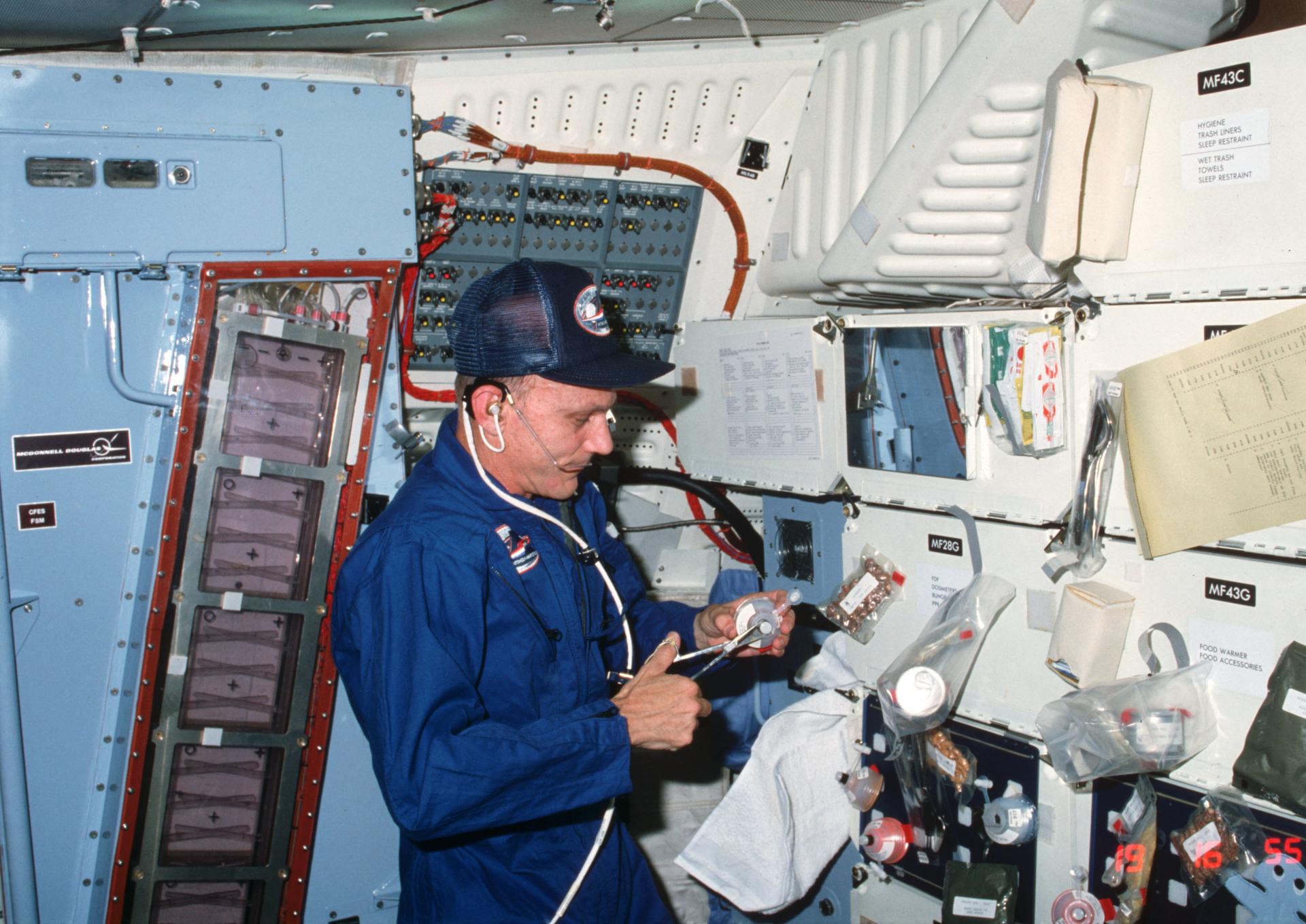 Astronaut Thomas K. Mattingly II, STS-4 crew commander, cuts open a drink container in preparation for a meal onboard Space Shuttle Columbia. Various packages of food and meal accessories are attached to locker doors. At far left edge of the frame is the tall payload called continuous flow electrophoresis experiment (CFES) system.