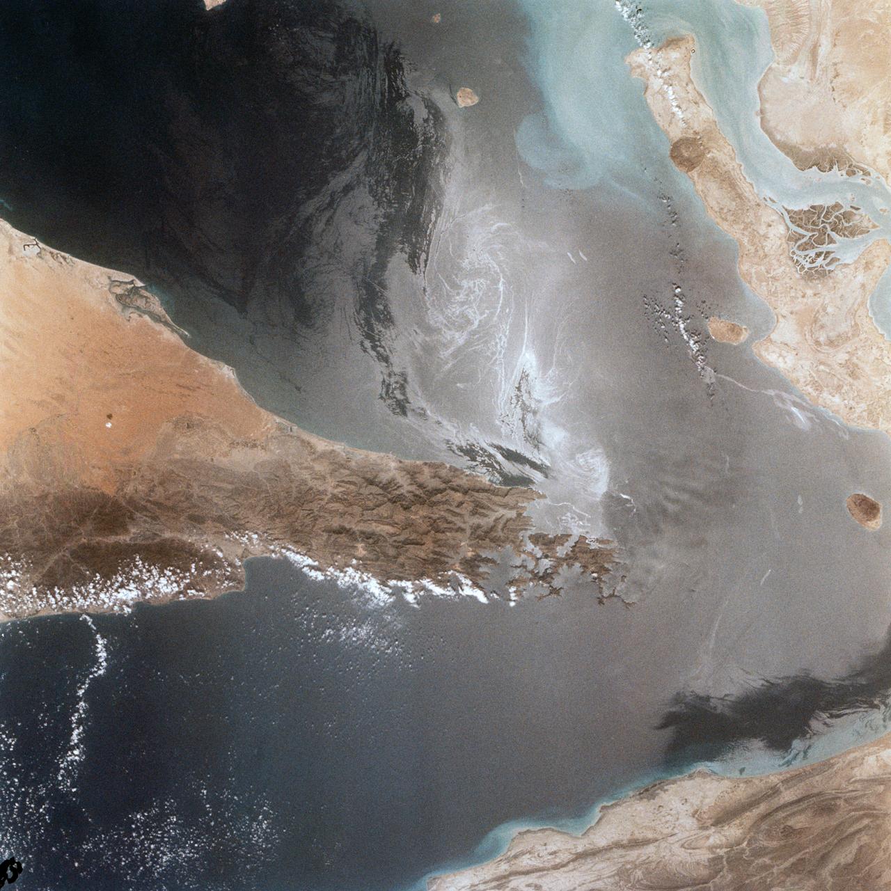 Portions of Oman, The United Arab Emirates and Iran are seen at the Strait of Hormuz (26.0N, 56.0E) in this view. A number of ship wakes can be seen in the area of the strait. Sunglint in the Persian Gulf to the northwest, accentuates the complex currents and oil slicks, seen as dark toned blue streaks on the surface. Qeshm Island, just off the coast of Iran, can be seen on the edge of the photo.