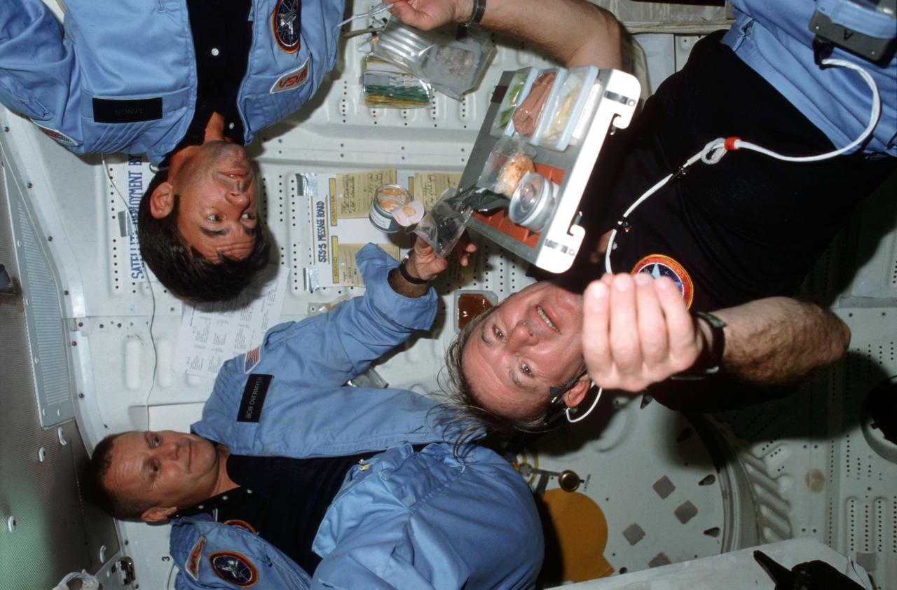STS005-06-210 (16 Nov. 1982) --- Astronaut Vance D. Brand (in dark blue shirt), STS-5 commander; Robert F. Overmyer (left), pilot; and William B. Lenoir, mission specialist, conduct microgravity experiments with food containers and meal tray assemblies in front of middeck port side wall and side hatch. Brand prepares to eat as meal tray assembly floats above his chest and Overmeyer and Lenoir look on. Sign on port side wall is labeled STS-5 message board. Photo credit: NASA
