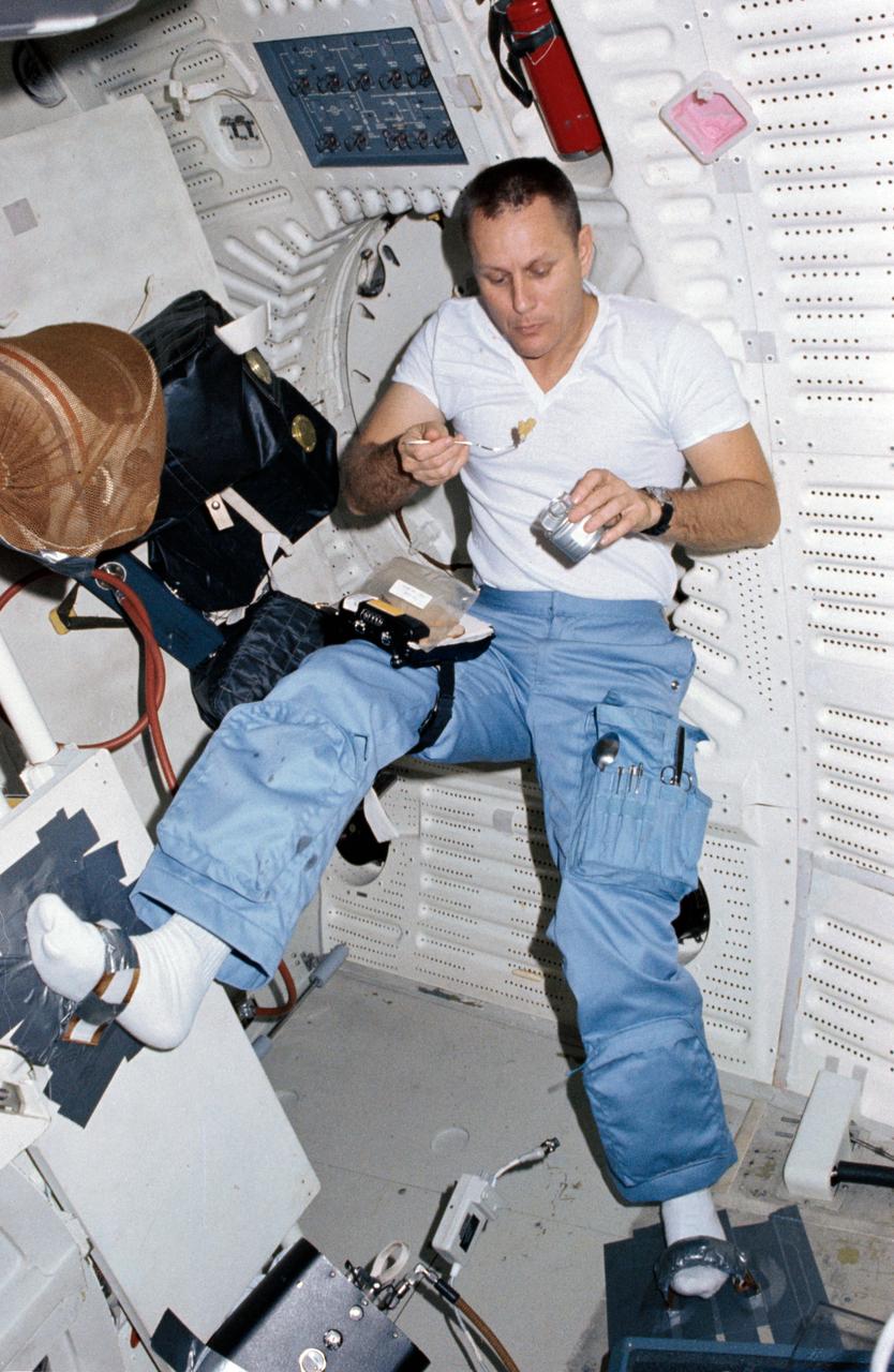 STS005-15-588 (13 Nov. 1982) --- Astronaut Robert F. Overmyer, STS-5 pilot, enjoys a meal from a jury-rigged set-up in the middeck area of the Earth-orbiting space shuttle Columbia.  He wears a T-shirt and the trouser portion of a multi-piece constant wear garment. His feet are positioned in recently-rigged foot restraints to avoid involuntary movement in the micro-gravity environment of space.   Behind Overmyer are components of the suit, including helmet, worn during landing and takeoff for shuttle flights. The trousers he is presently wearing are part of that attire. Photo credit: NASA
