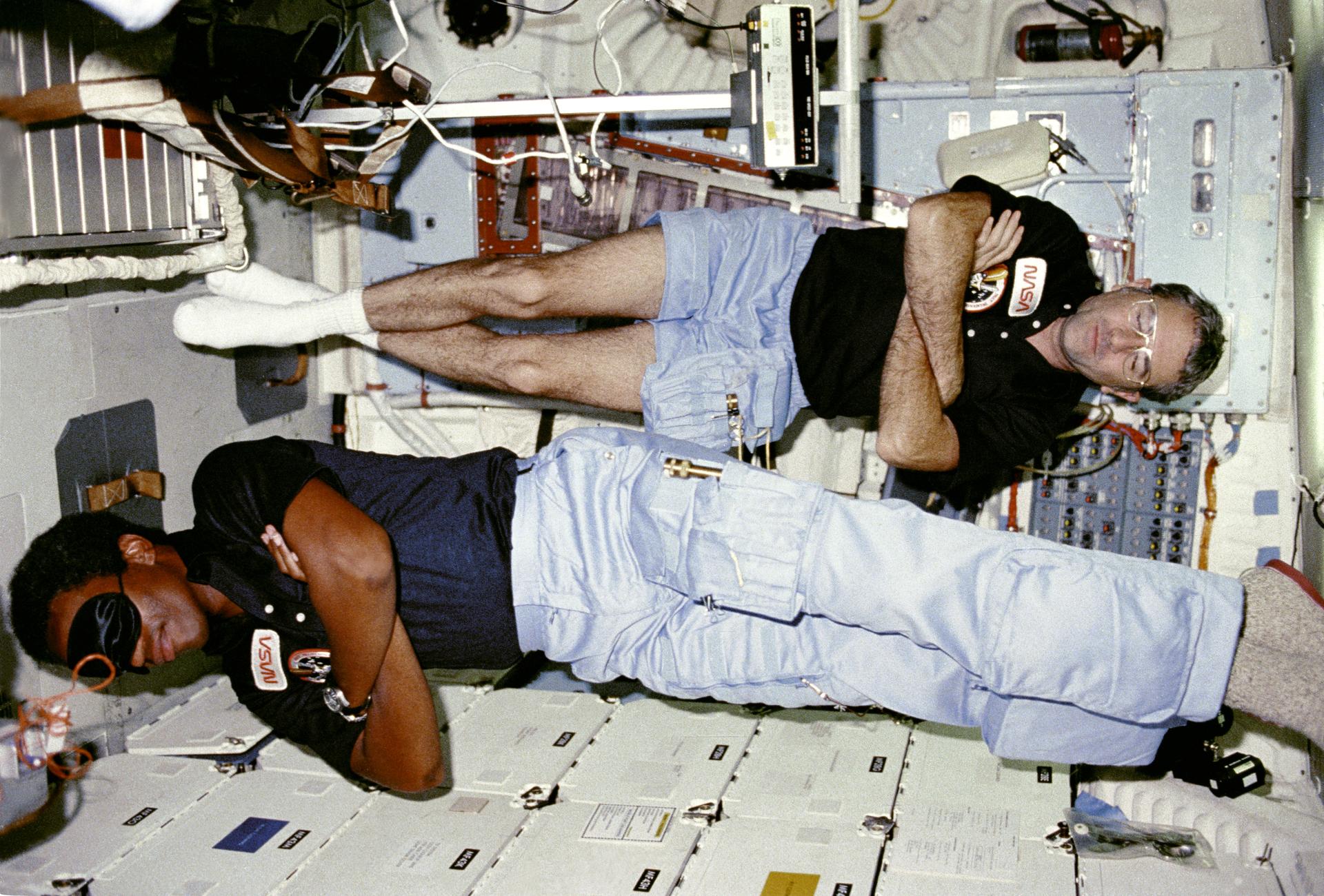 Richard Truly and Guy Bluford float with their arms crossed inside the Space Shuttle as they sleep for a bit during the STS-8 mission.