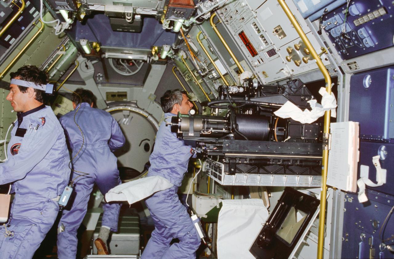STS009-03-093 (28 Nov-8 Dec 1983) --- A mission specialist and two payload specialists busy themselves in the Spacelab 1 module aboard the Earth orbiting Space Shuttle Columbia.  Left to right are Payload Specialist Robert A. R. Parker.  Parker is partially obscured by a deployed instrument of the fluid physics module at the materials sciences double rack.  Merbold, a physicist from Max-Planck Institute in the Federal Republic of Germany, wears a head band-like device and a recorder as part of an overall effort to learn more about space adaptation.  Both Space lab 1 payload specialists wore the devices during most of their waking hours on this 10-day flight.  The frame was exposed with a 35mm camera.