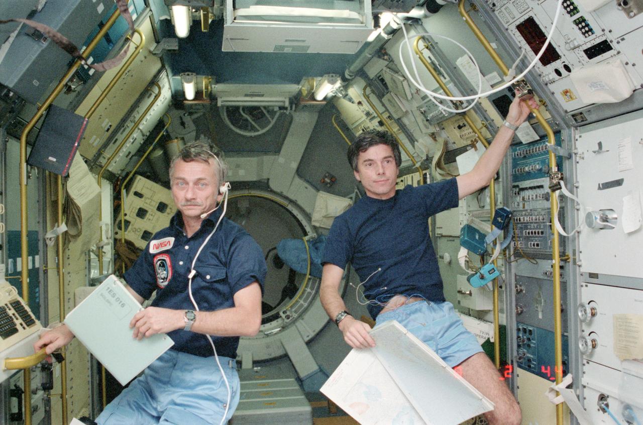 STS009-123-340 (28 Nov 1983) --- Astronaut Owen K. Garriott, STS-9 mission specialist, left, and Ulf Merbold, payload specialist, take a break from monitoring experimentation aboard Spacelab to be photographed. Dr. Garriott, holds in his left hand a data/log book for the solar spectrum experiment. Dr. Merbold, holds a map in his left hand for the monitoring of ground objectives of the metric camera.