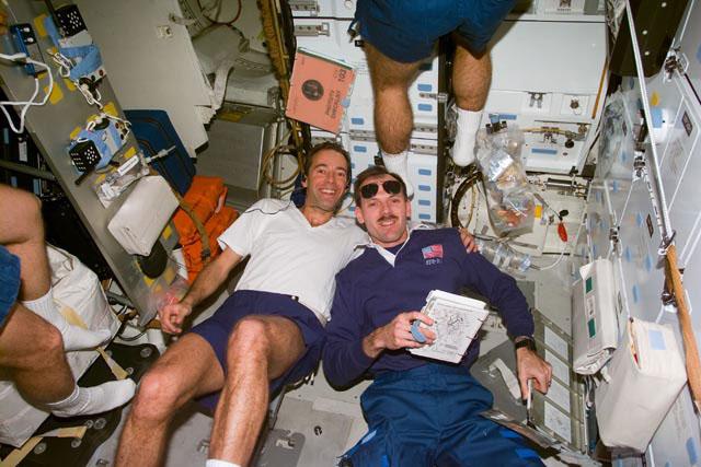 S103-E-5194 (21 December 1999) --- Astronaut Jean-Francois Clervoy (left), mission specialist representing the European Space Agency (ESA), works out on an ergometer device;  and astronaut Steven L. Smith, payload commander, fetches a reference manual.   Two unidentified STS-103 crew mates are busy at various tasks, partially out of the frame. This photo was taken with an electronic still camera (ESC) at 11:08:38 GMT, Dec. 21, 1999.