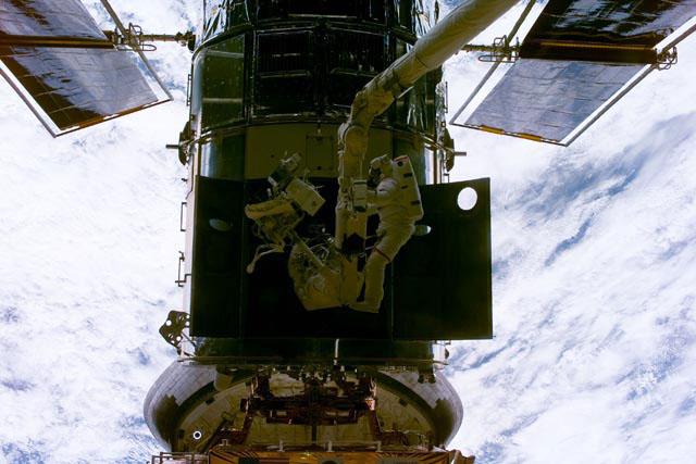 STS-103 mission specialist John M. Grunsfeld (attached to a workstation on the RMS arm) and payload commander Steven L. Smith (free-floating) perform a changeout of the Rate Sensor Units (RSU) in one of the bays of -V3 plane of the Hubble Space Telescope (HST). This repair was performed during the first of three extravehicular activities (EVAs) of the mission. Grunsfeld is distinguished by having no marks on his EMU and Smith is distinguished by the red strip on the pants of his EMU.