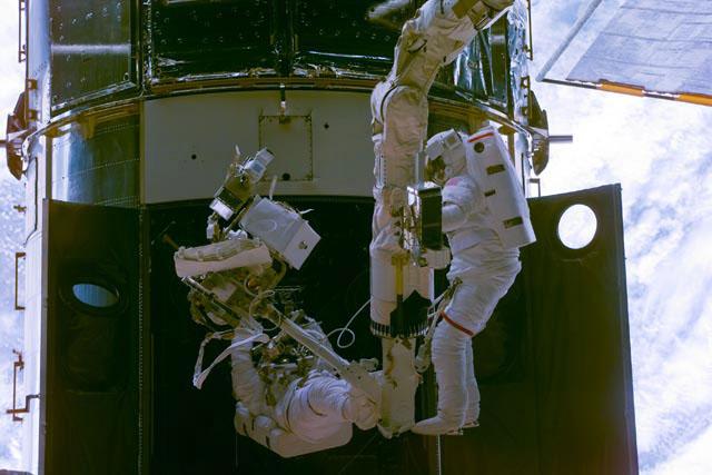 S103-E-5204 --- Astronauts Steven L. Smith (right), payload commander, and  John M. Grunsfeld use the Canadian-built remote manipulator system (RMS) robot arm to perform servicing tasks on the temporarily-captured Hubble Space Telescope (HST).  The photo was taken with an electronic still camera.