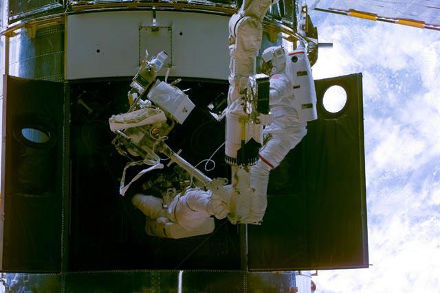 STS-103 mission specialist John M. Grunsfeld (attached to a workstation on the RMS arm) and payload commander Steven L. Smith (free-floating) perform a changeout of the Rate Sensor Units (RSU) in one of the bays of -V3 plane of the Hubble Space Telescope (HST). This repair was performed during the first of three extravehicular activities (EVAs) of the mission. Grunsfeld is distinguished by having no marks on his EMU and Smith is distinguished by the red strip on the pants of his EMU.