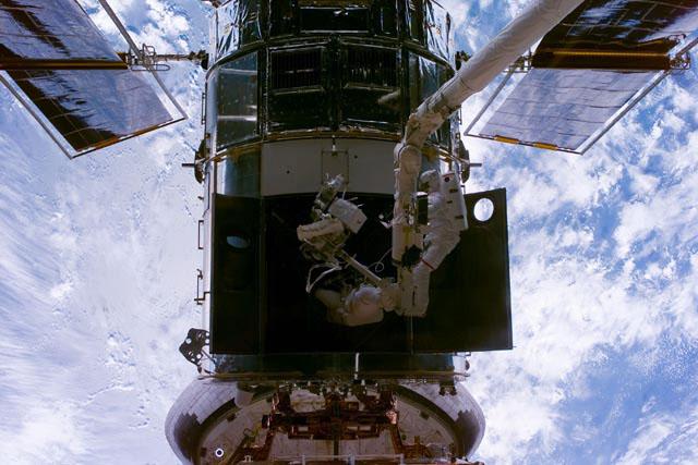 S103-E-5206 --- Astronauts Steven L. Smith (right), payload commander, and  John M. Grunsfeld use the Canadian-built remote manipulator system (RMS) robot arm to perform servicing tasks on the temporarily-captured Hubble Space Telescope (HST).  The photo was taken with an electronic still camera (ESC).