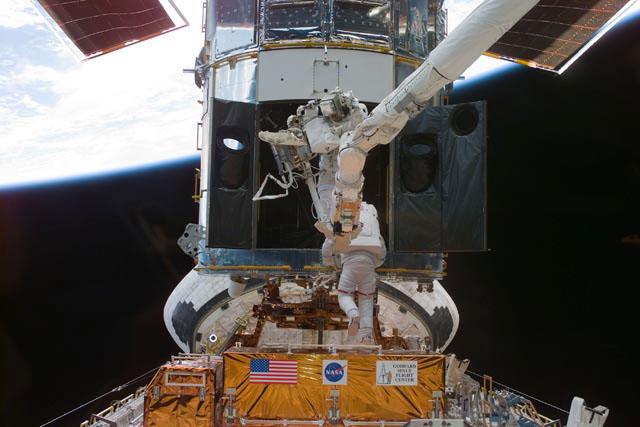 S103-E-5208 --- Astronauts Steven L. Smith (bottom), payload commander, and  John M. Grunsfeld, mission specialist, perform servicing tasks on the temporarily-captured Hubble Space Telescope (HST). Grunsfeld is on a foot restraint connected to Discovery's remote manipulator system (RMS) robot arm. Smith, making his second servicing visit to HST, is using handrails on the telescope. The photo was taken with an electronic still camera (ESC).