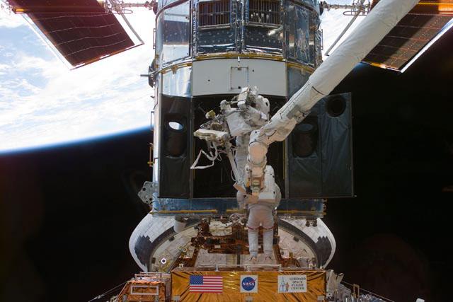 S103-E-5209 --- Astronauts Steven L. Smith (bottom), payload commander, and  John M. Grunsfeld, mission specialist, perform servicing tasks on the temporarily-captured Hubble Space Telescope (HST). Grunsfeld is on a foot restraint connected to Discovery's remote manipulator system (RMS) robot arm. Smith, making his second servicing visit to HST, is using handrails on the telescope. The photo was taken with an electronic still camera (ESC).