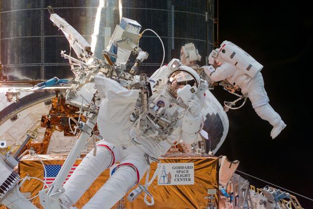 S103-E-5347 (24 December 1999) --- Astronaut Steven L. Smith, payload commander, prepares to use a 35mm camera during the final space walk of the STS-103 mission.  Smith is standing  on a foot restraint connected to the end of Discovery's remote manipulator system (RMS) robot arm. Astronaut John M. Grunsfeld, mission specialist, translates along a handrail system on  the Hubble Space Telescope (HST) in the background.