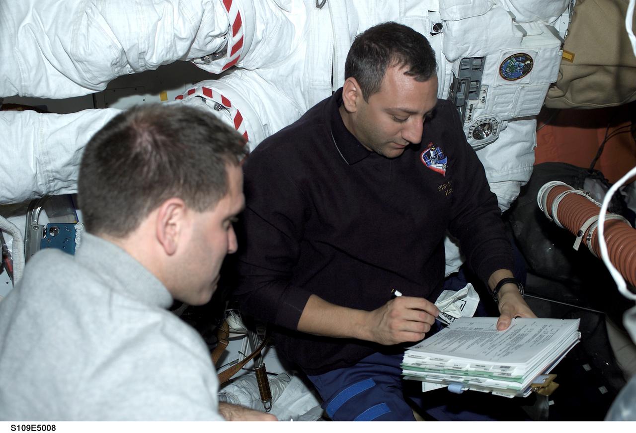 STS109-E-5008 (3 March 2002) --- On the mid deck of the Space Shuttle Columbia, astronauts John M. Grunsfeld (foreground), payload commander, and Michael J. Massimino, mission specialist, go over a checklist concerning the next few days' scheduled space walks. Massimino's extravehicular mobility unit (EMU) space suit, which will be called into duty for the second day of extravehicular activity (EVA), is in the background. The image was recorded with a digital still camera.