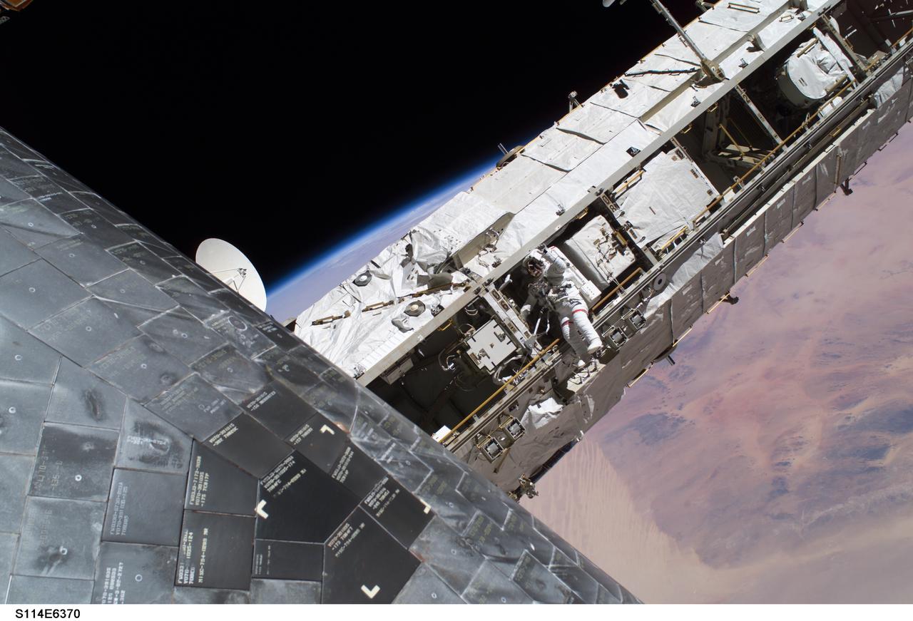 S114-E-6370 (3 August 2005) --- While perched on a Space Station truss, astronaut Soichi Noguchi, STS-114 mission specialist representing Japan Aerospace Exploration Agency (JAXA), acts as observer and communication relay station between fellow spacewalker Stephen K. Robinson (out of frame) and astronaut Andrew S. W. Thomas aboard Space Shuttle Discovery during the mission’s third session of extravehicular activities (EVA). A portion of the thermal protection tiles on Discovery’s underside is visible at lower left. The image was photographed by Robinson using a digital camera.
