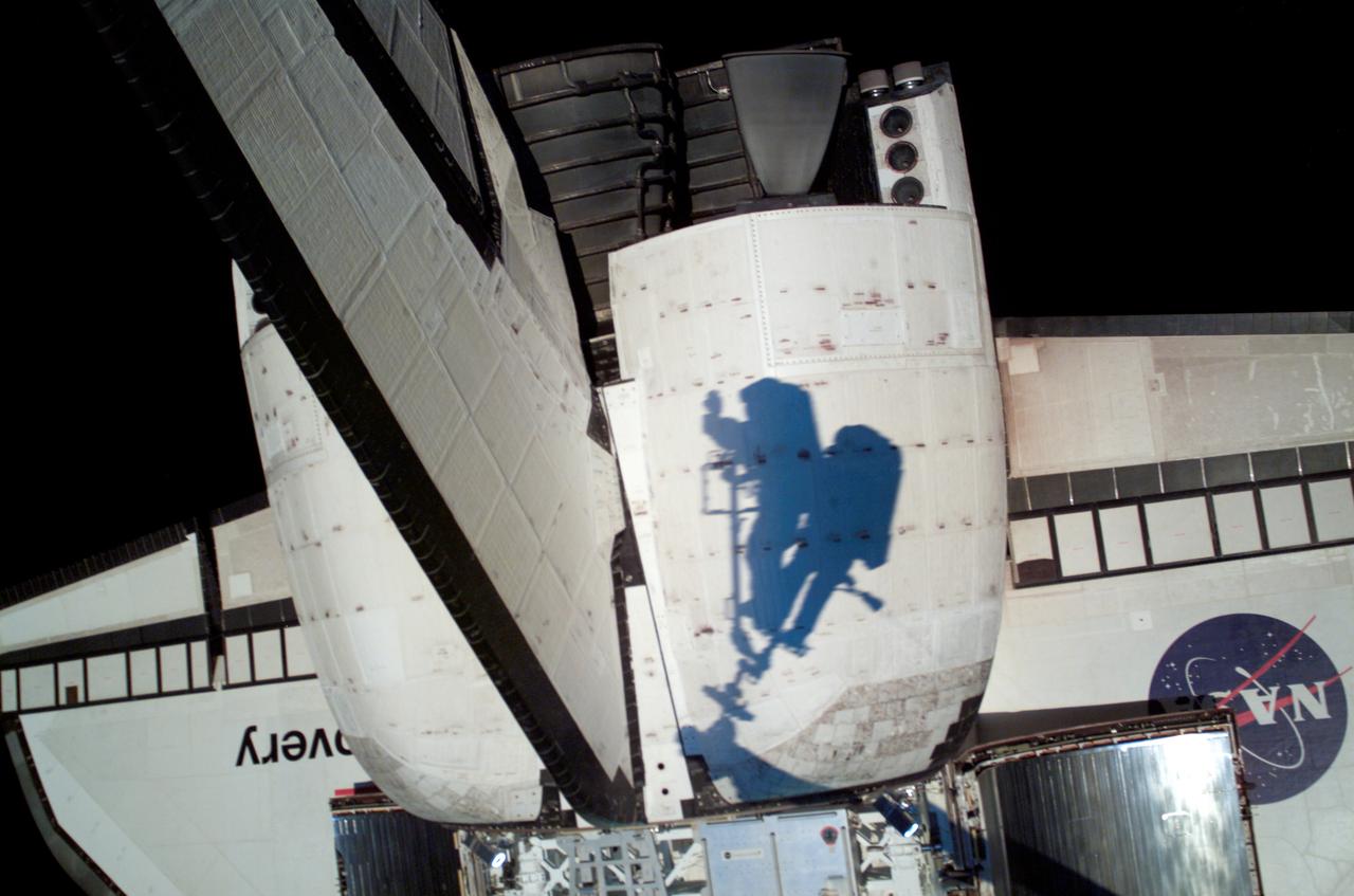 S121-E-06388 (8 July 2006) --- The shadows of astronauts Piers J. Sellers and Michael E. Fossum, two STS-121 mission specialists who are anchored to the Space Shuttle Discovery's Remote Manipulator System/Orbiter Boom Sensor System (RMS/OBSS) foot restraint, are visible against the port pod for the Orbital Maneuvering System on the shuttle. Photo credit: NASA