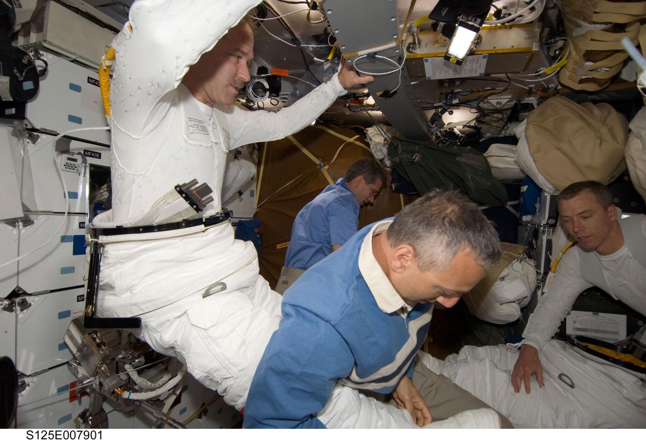S125-E-007901 (16 May 2009) --- Astronaut John Grunsfeld gets help from astronaut Mike Massimino, both STS-125 mission specialists, in the donning of his Extravehicular Mobility Unit (EMU) spacesuit on the middeck of the Earth-orbiting Space Shuttle Atlantis in preparation for the mission?s third session of extravehicular activity (EVA). Astronaut Andrew Feustel is at right and Michael Good works in the background.