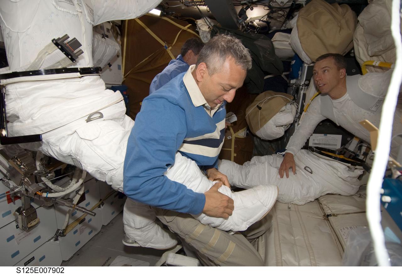 S125-E-007902 (16 May 2009) --- Astronaut Mike Massimino, STS-125 mission specialist, assists astronaut John Grunsfeld (partially out of frame), mission specialist, in the donning of his Extravehicular Mobility Unit (EMU) spacesuit on the middeck of the Earth-orbiting Space Shuttle Atlantis in preparation for the mission?s third session of extravehicular activity (EVA). Astronaut Andrew Feustel, mission specialist, is at right.