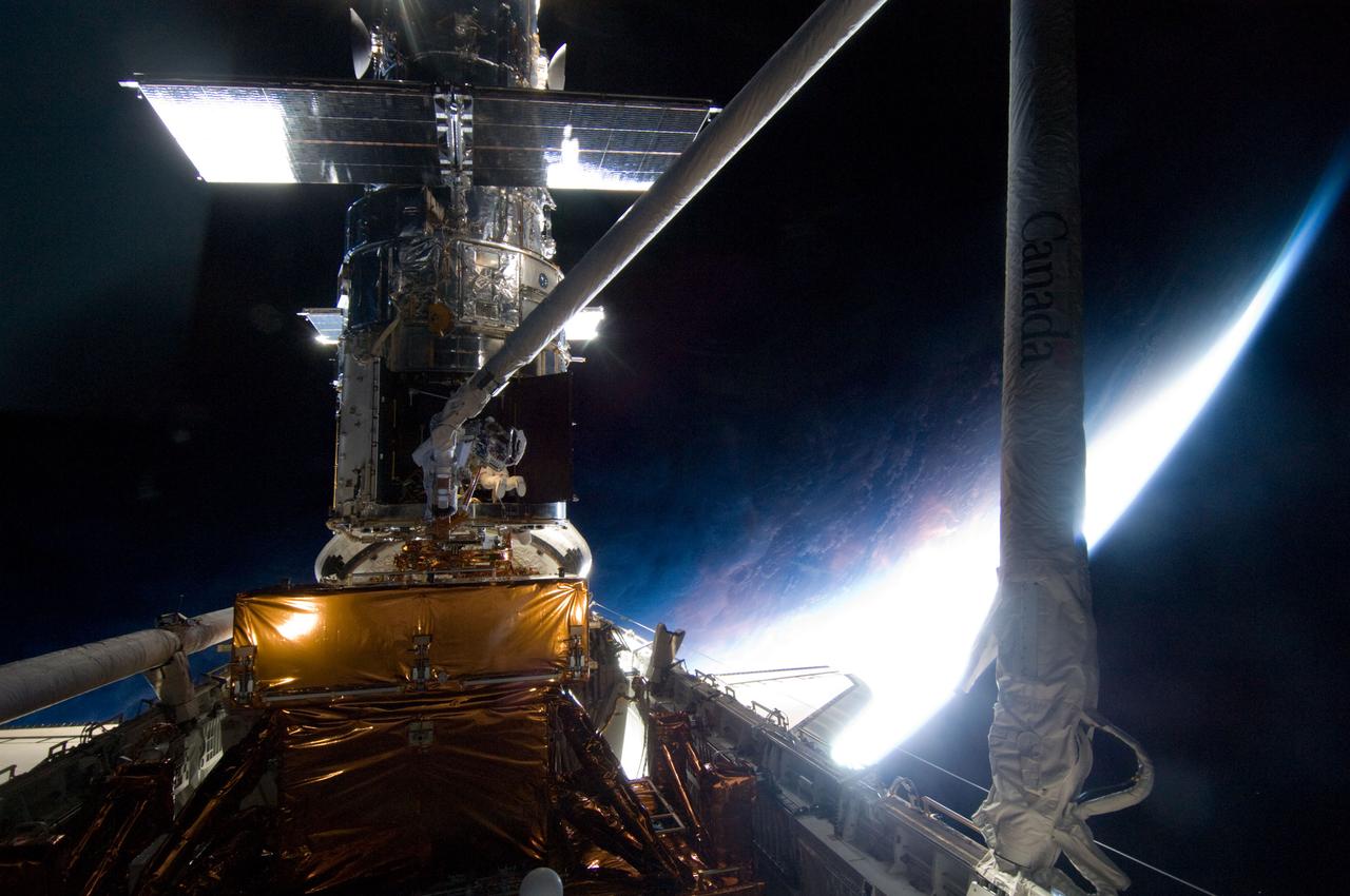 S125-E-009194  (17 May 2009) --- With a mostly dark home planet behind him, astronaut Michael Good, STS-125 mission specialist, rides Atlantis' remote manipulator system arm to the exact position he needs to be to continue work on the Hubble Space Telescope.  Astronaut Mike Massimino, who shared two spacewalks with Good during the last week, is out of frame.