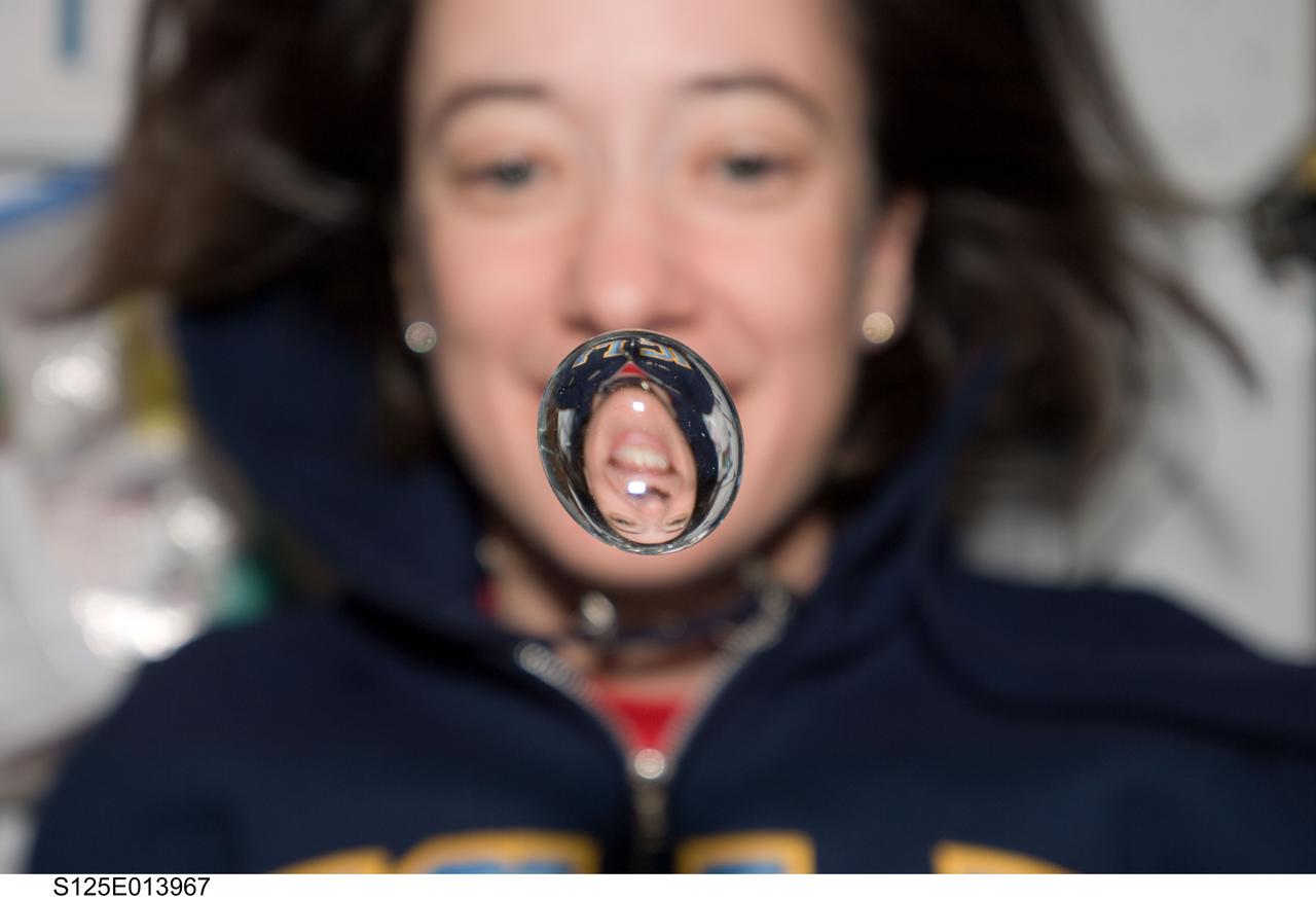 View of STS-125 Mission Specialist 2 (MS2) Megan McArthur posing for a photo on the middeck. A water bubble, showing a reflection of McArthur, floats in front of her.