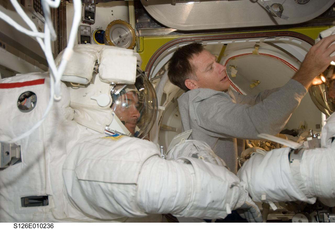 S126-E-010236 (24 Nov. 2008) --- Astronauts Shane Kimbrough and Steve Bowen (right, mostly out of frame), both STS-126 mission specialists, attired in their Extravehicular Mobility Unit (EMU) spacesuits, are pictured in the Quest Airlock of the International Space Station as the mission's fourth session of extravehicular activity (EVA) draws to a close. Astronaut Chris Ferguson, commander, assists with the doffing of the spacesuits.