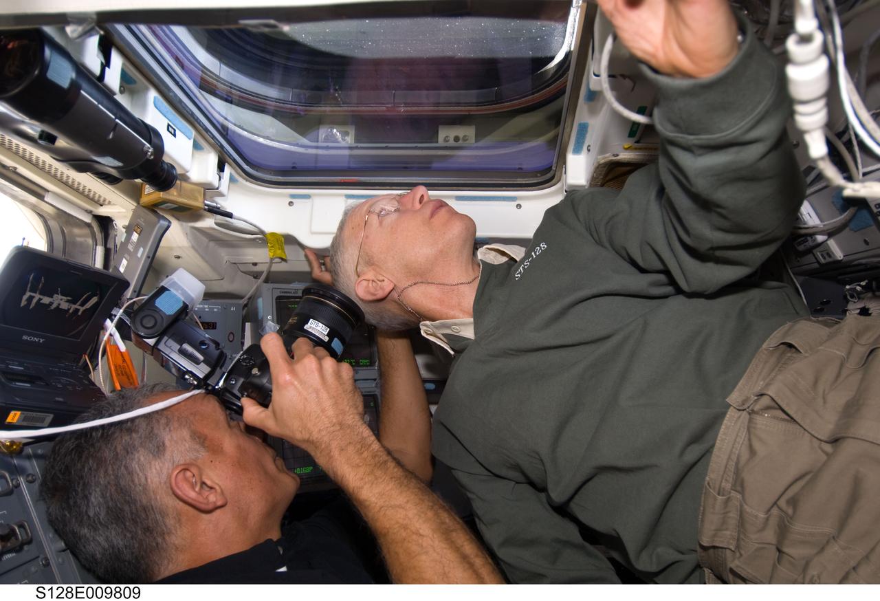S128-E-009809 (8 Sept. 2009) --- NASA astronauts John “Danny” Olivas (left) and Patrick Forrester, both STS-128 mission specialists, are pictured on the aft flight deck of Space Shuttle Discovery during flight day 12 activities. Olivas is using a still camera at an overhead window to photograph the International Space Station after the undocking of the two spacecraft.