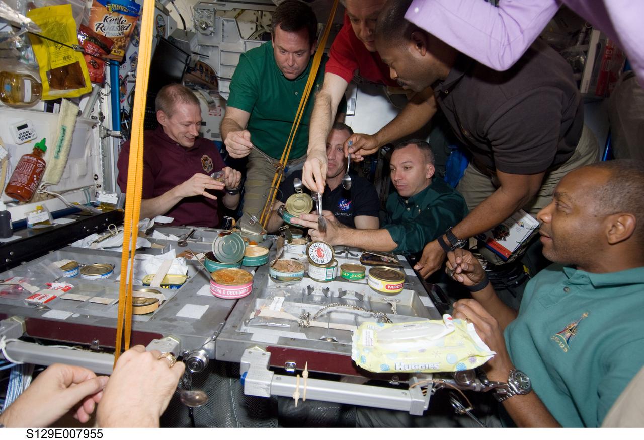 S129-E-007955 (23 Nov. 2009) --- Expedition 21 and STS-129 crew members gather for a meal at the galley in the Unity node of the International Space Station while space shuttle Atlantis remains docked with the station. Pictured are European Space Agency astronaut Frank De Winne, Expedition 21 commander; NASA astronauts Charles O. Hobaugh, STS-129 commander; Mike Foreman, Randy Bresnik, Leland Melvin and Robert L. Satcher Jr., all STS-129 mission specialists; along with Canadian Space Agency astronaut Robert Thirsk, Expedition 21 flight engineer.