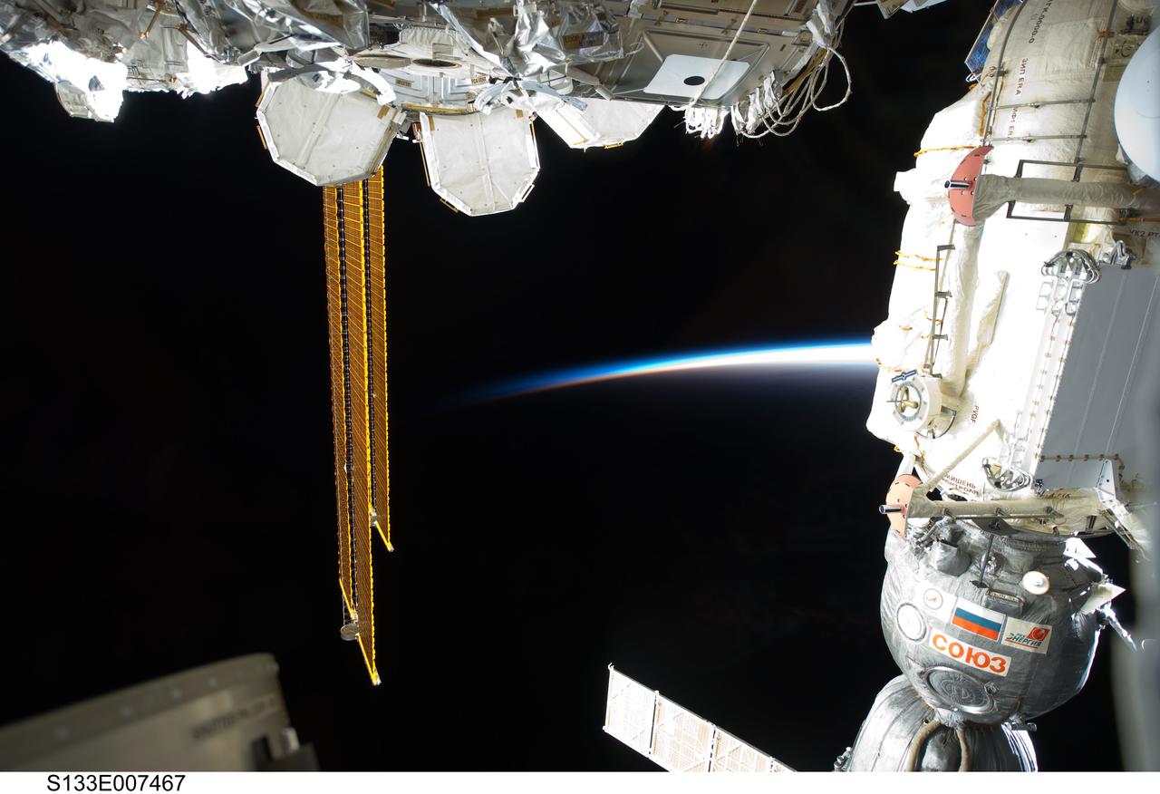 S133-E-007467 (1 March 2011) --- Backdropped by the thin line of Earth’s atmosphere and the blackness of space, a docked Russian Soyuz spacecraft (right), a portion of the International Space Station’s Quest airlock and solar array panels are featured in this image photographed by an STS-133 crew member while space shuttle Discovery remains docked with the station. Photo credit: NASA or National Aeronautics and Space Administration