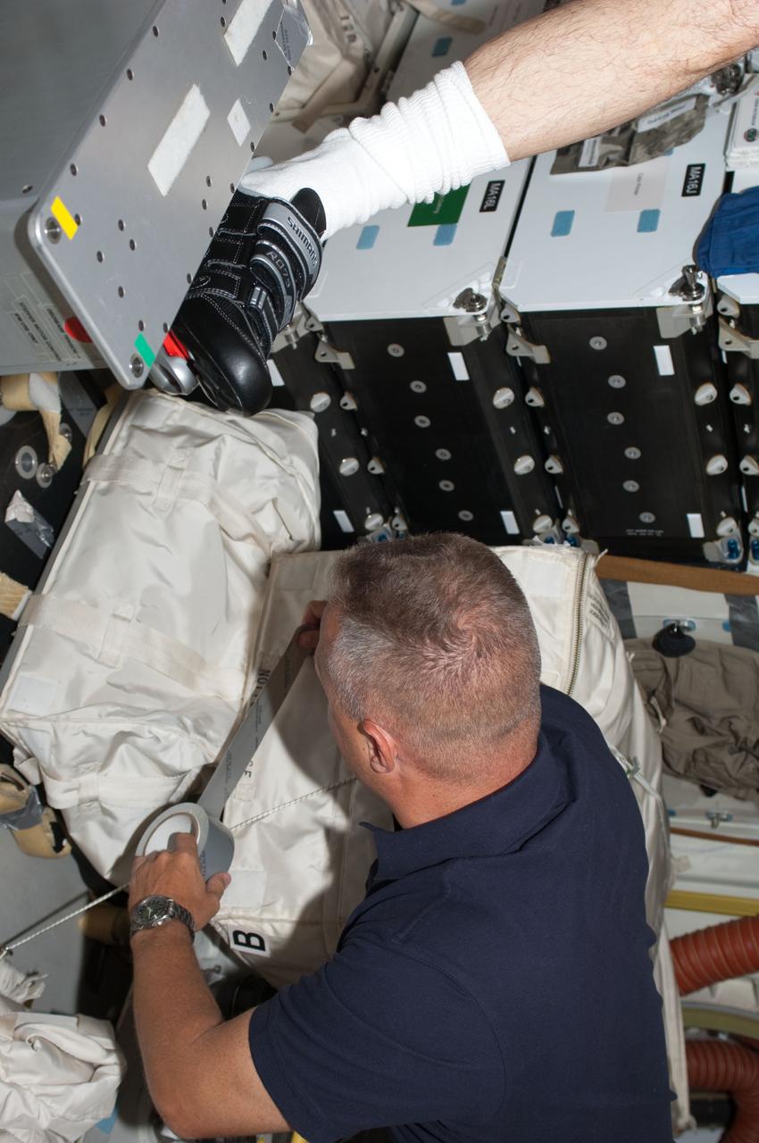 S135-E-008110 (13 July 2011) --- NASA astronaut Doug Hurley, STS-135 pilot, tapes up a supply bag on the middeck of the space shuttle Atlantis while the spacecraft was docked with the International Space Station. Most of the ten crewmembers were involved in transfer operations. In this scene, a crewmate, whose left foot is visible, works out on the ergometer device at top. Photo credit: NASA