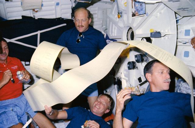 NASA image: STS-26 crewmembers eat on middeck as TAGS printout drifts among them