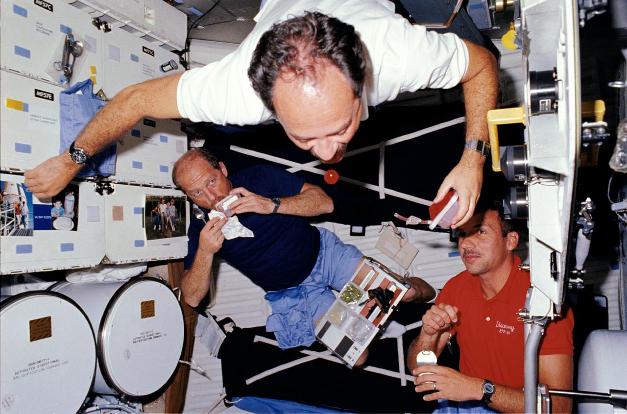 STS026-06-018 (29 Sept. - 3 Oct. 1988) --- Astronaut John M. Lounge, STS-26 mission specialist (MS), using a beverage container, experiments with microgravity as Commander Frederick H. Hauck (left) and MS David C. Hilmers (right) look on. Lounge freefloats as he closes in on a sphere of the red liquid drifting in front of his mouth. Hauck holds a spoon while sipping from a beverage container as he balances a meal tray assembly on his thighs. Hilmers, partially blocked by the open airlock hatch and holding a spoon and a can of food, pauses to watch the experiment. Automated Directional Solidification Furnace (ADSF) and forward middeck lockers appear on Lounge's right.