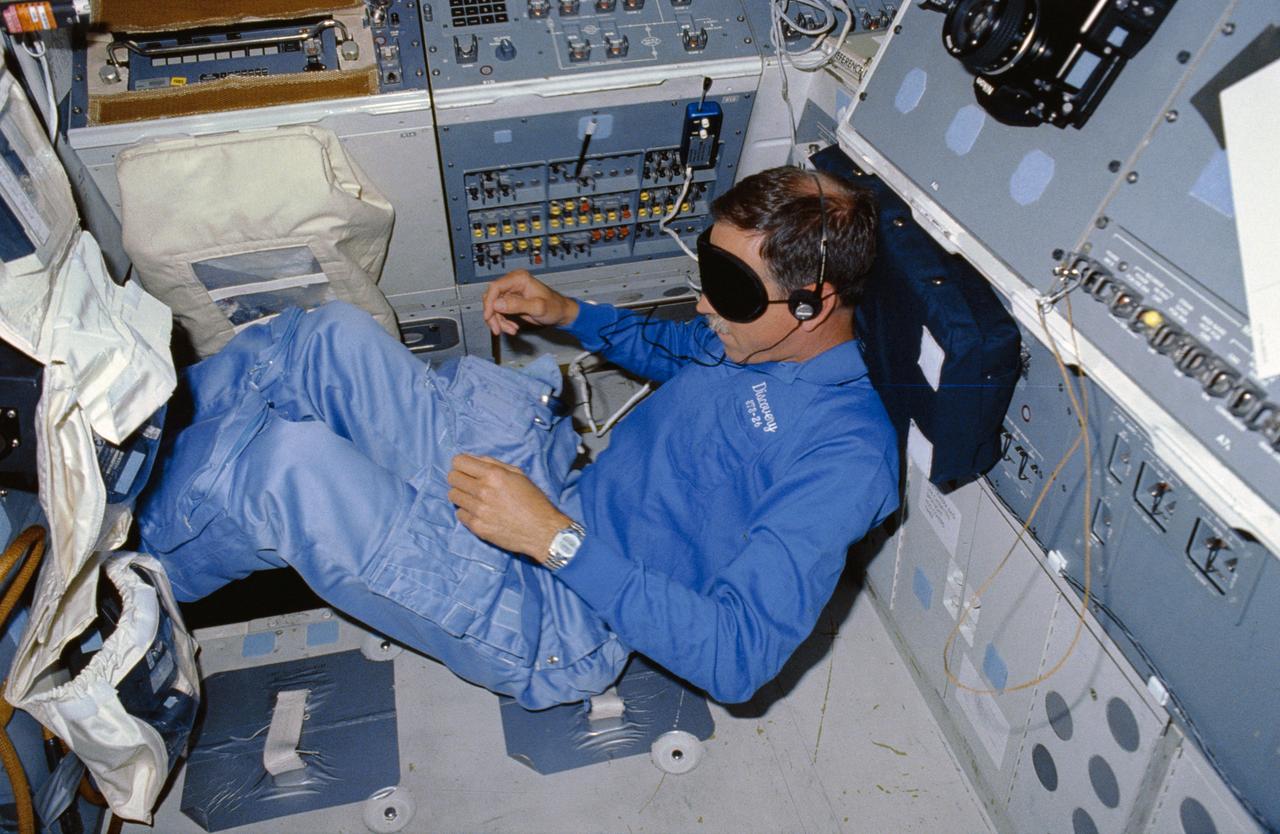 STS026-09-021 (3 Oct 1988) --- Astronaut Richard O. Covey, STS-26 pilot, wearing sleep mask (blindfold) and a headset, props his feet under the pilots seat and rests his head and back on the aft flight deck on orbit station panels while he sleeps. At Covey's right are the mission station control panels.