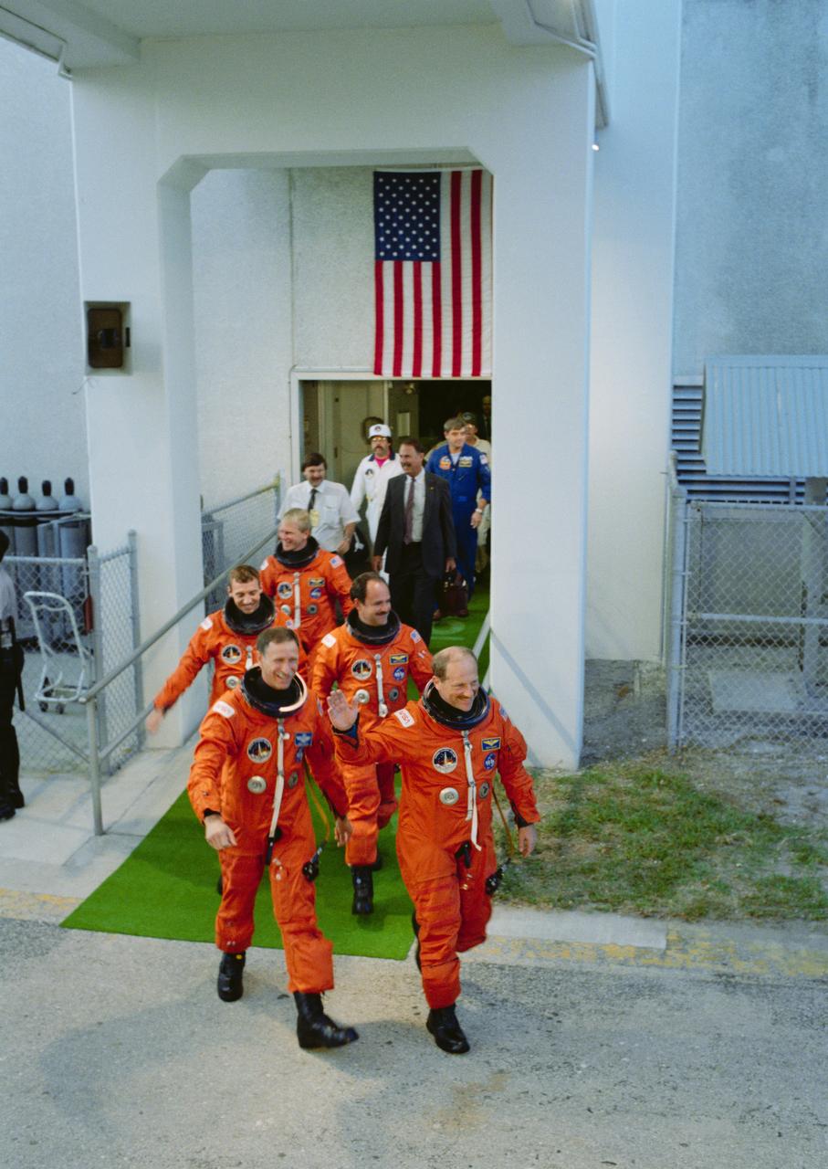 STS-26 crewmembers, suited in launch and entry suits (LESs), leave the Kennedy Space Center (KSC) Operations and Checkout (O&C) Building on their way to the launch complex (LC) pad 39B where they will board Discovery, Orbiter Vehicle (OV) 103. Commander Frederick H. Hauck, waving to spectators, is followed by Pilot Richard O. Covey, Mission Specialist (MS) John M. Lounge, MS David C. Hilmers, and MS George D. Nelson. Others accompanying the crew are JSC Flight Crew Operations Directorate (FCOD) Director Donald R. Puddy, Astronaut Office Chief Daniel C. Brandenstein, and a Crew Training Officer Richard W. Nygren.