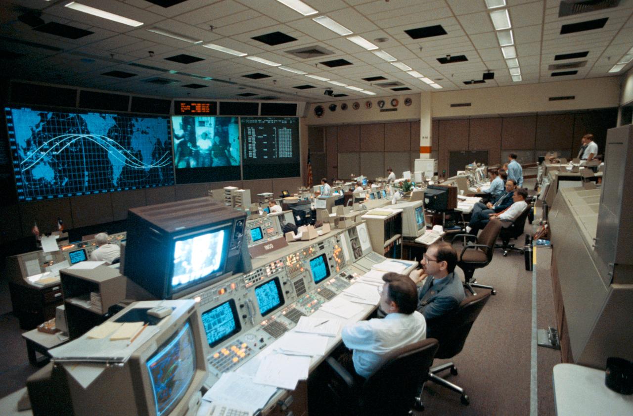 Flight controllers in JSC's Mission Control Center (MCC) Bldg 30 flight control room (FCR) listen to a presentation by STS-26 crewmembers on the fourth day of Discovery's, Orbiter Vehicle (OV) 103's, orbital mission. Instrumentation and Communications Officers (INCOs) Harold Black (left foreground) and John F. Muratore and other controllers view a television (TV) transmission of the crew on a screen in front of the FCR as each member relates some inner feelings while paying tribute to the 51L Challenger crew.