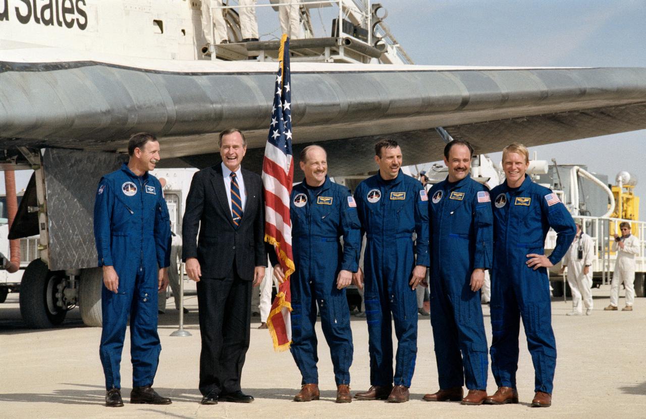 STS026-S-169 (3 Oct 1988) --- The five STS 26 crewmembers pose with Vice President George Bush after completing a successful four-day mission aboard the Space Shuttle Discovery (background). Pictured, from left to right, are astronaut Richard O. Covey, pilot; Vice President Bush; and astronauts Frederick H. (Rick) Hauck, mission commander; and David C. Hilmers, John M. (Mike) Lounge, and George D. Nelson, all mission specialists.