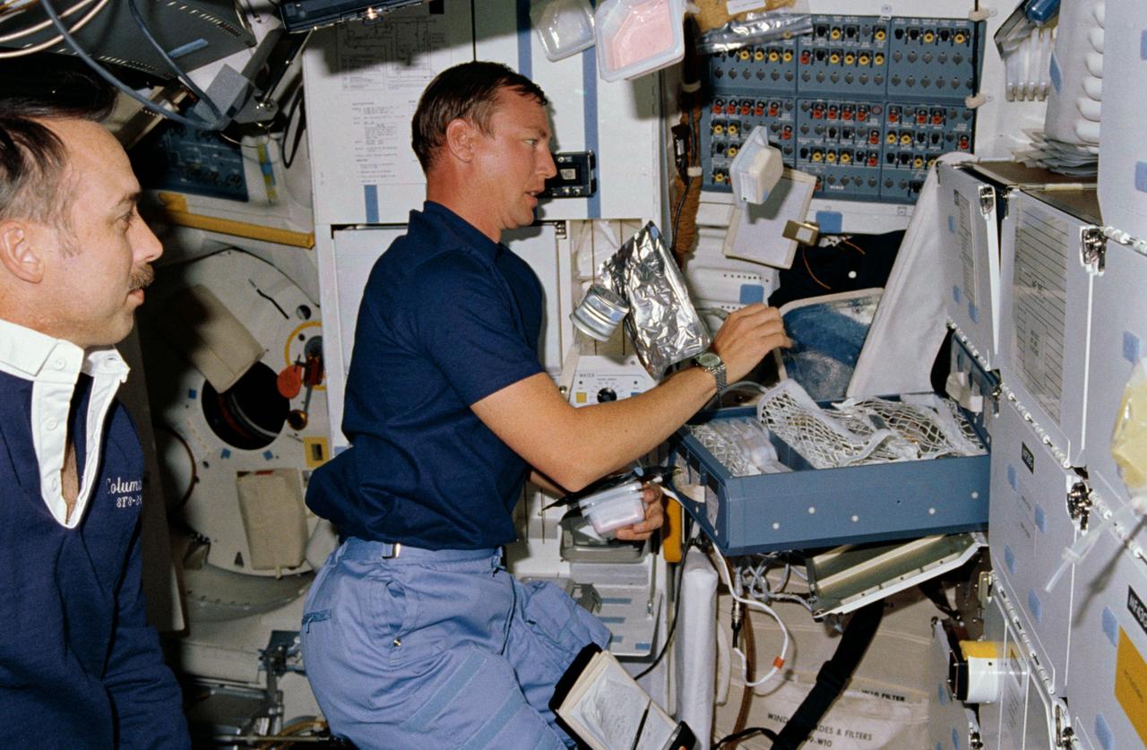 STS028-13-013 (August 1989) --- Astronaut Mark N. Brown, STS-28 mission specialist, assembles various components of a meal at the stowage locker area of Columbia's middeck, as James C. Adamson, mission specialist, looks on.