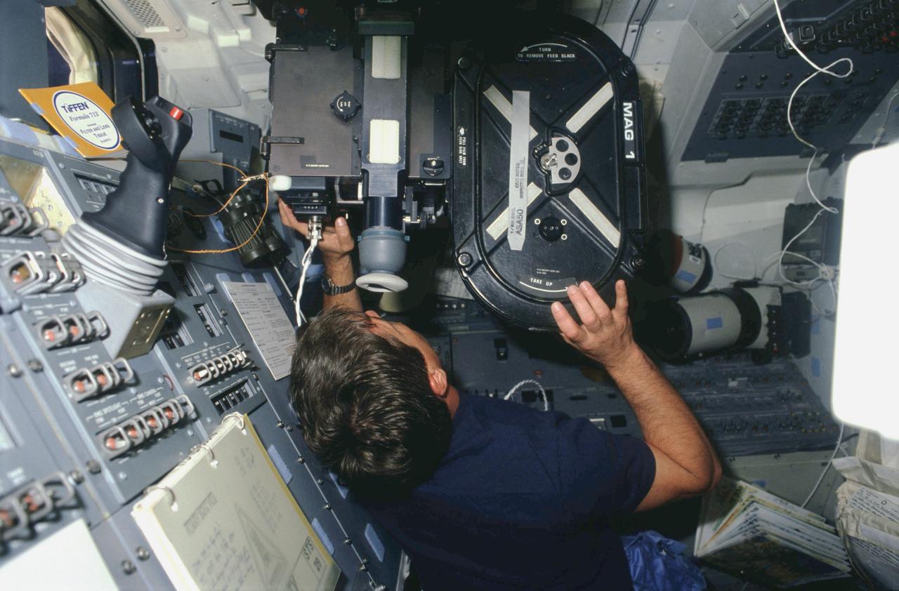 Pilot John E. Blaha balances and points IMAX motion picture camera out aft flight deck overhead window to film Earth's surface below. Blaha is surrounded by onorbit station control panels in foreground, payload station in background, and forward overhead panels above. Handbook titled "TiFFEN Formula 712" and checklists are velcroed to onorbit station panels. An open atlas freefloats just below Blaha's right elbow.
